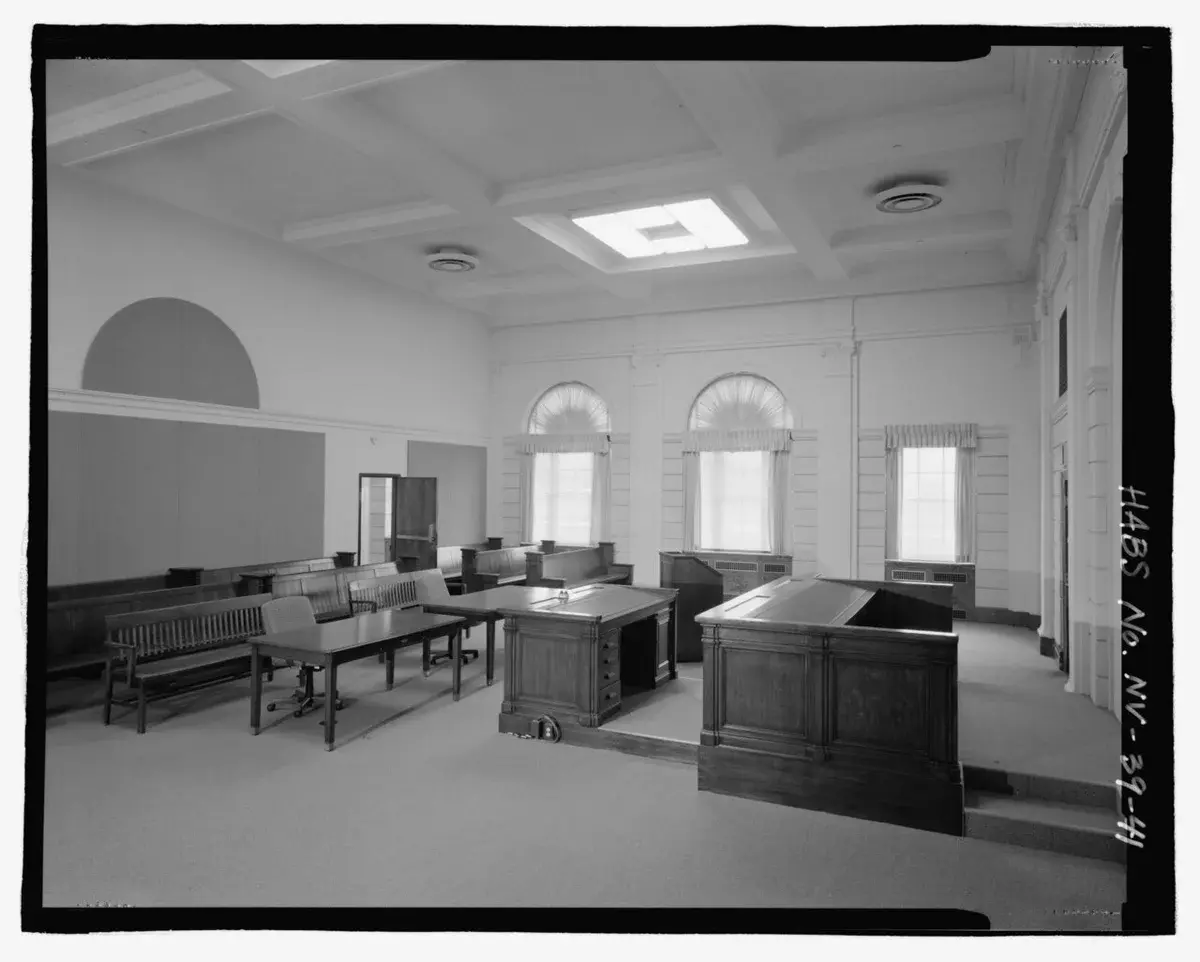 Black-and-white photo of a historic courtroom interior with benches, tables, and arched windows.