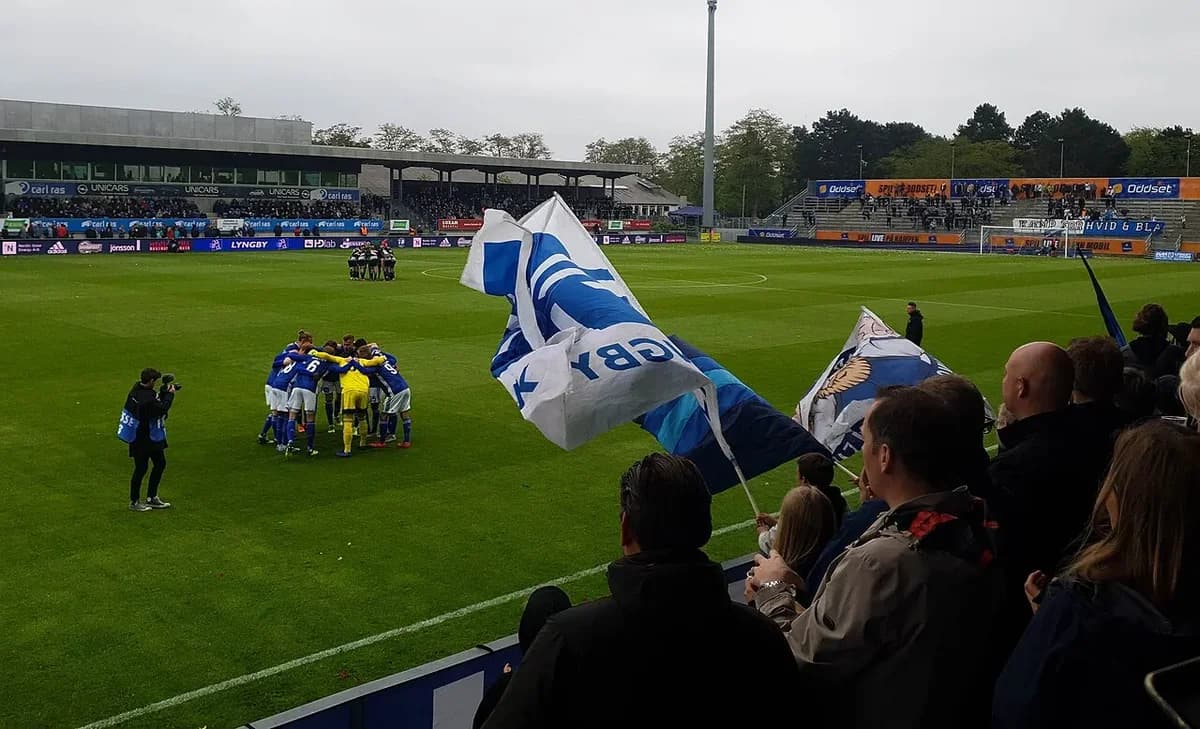 Football teams huddled on the pitch with fans waving flags at a stadium