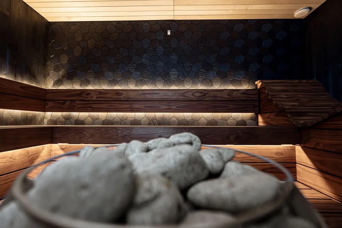 A modern sauna interior with wood benches, textured dark wall panels, and a basket of stones in the foreground.