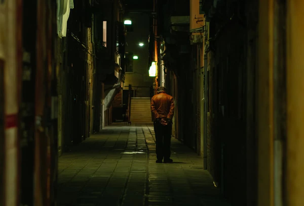 An older man walking alone down a narrow alley at night.