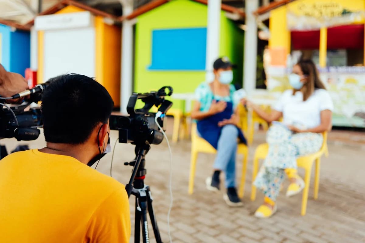 A film crew records an outdoor interview with two masked women.